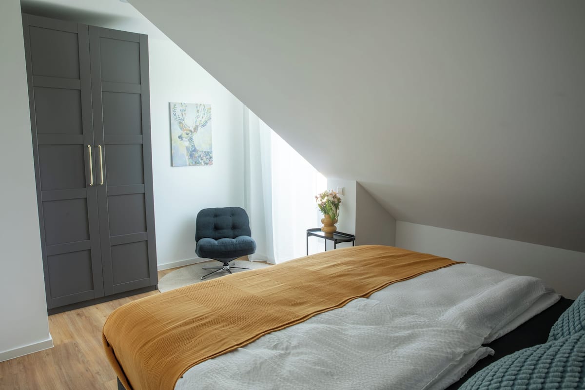 Un dormitorio con una cama grande, un armario oscuro, una silla y una mesita con un florero.