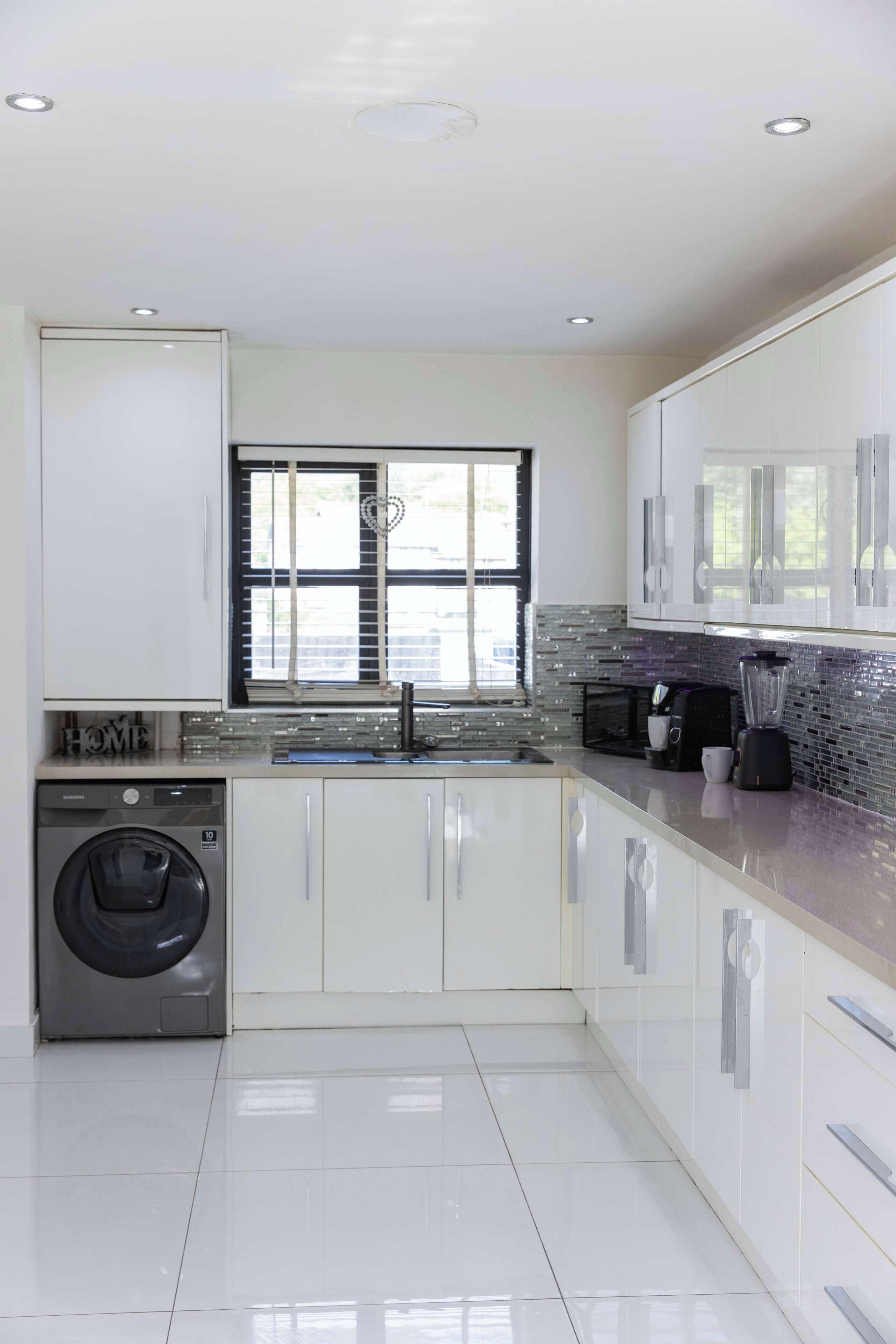 Una cocina moderna con armarios blancos, lavadora integrada, ventana con persianas y salpicadero de azulejos.