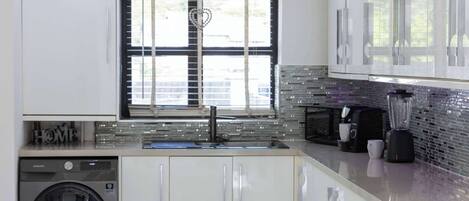 Una cocina moderna con armarios blancos, lavadora integrada, ventana con persianas y salpicadero de azulejos.