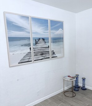 A room with a white wall, a wooden floor, and a framed picture of a pier extending into the sea. - Sailboat Bend House | Beach Days & Sunset Nights (Fort Lauderdale)