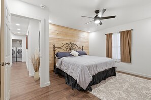 A bedroom with a bed, a ceiling fan, a wooden wall, and a view of a laundry area.