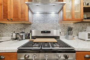 A kitchen with a gas stove, stainless steel appliances, wooden cabinets, and a hexagonal tile backsplash.
