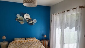 Een slaapkamer met een blauwe wand, een bed met een bedovertrek met dessin, decoratieve wandkunst, een raam met doorschijnende gordijnen en een hanglamp.