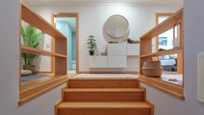 Un escalier avec des marches en bois menant à un palier, un mur blanc orné d’un miroir rond et une fenêtre donnant sur un lit.