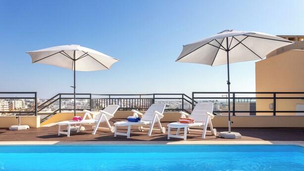 Une piscine sur le toit, agrémentée de chaises longues blanches et de parasols, offrant une vue panoramique sur la ville.