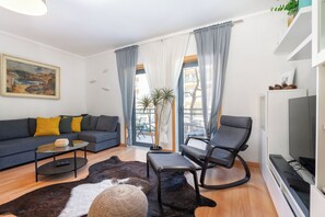 Ein modernes Wohnzimmer mit einer Couch, einem Couchtisch, einem Sessel, einem Fernseher und einem großen Fenster mit Vorhängen.