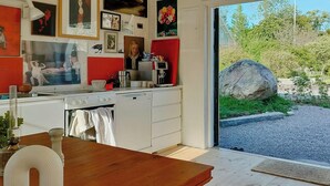 A modern kitchen with white cabinets, a wooden dining table, and a large window overlooking a gravel path and greenery. - Converted 1800s stable near Kalmarsund (Söderåkra)