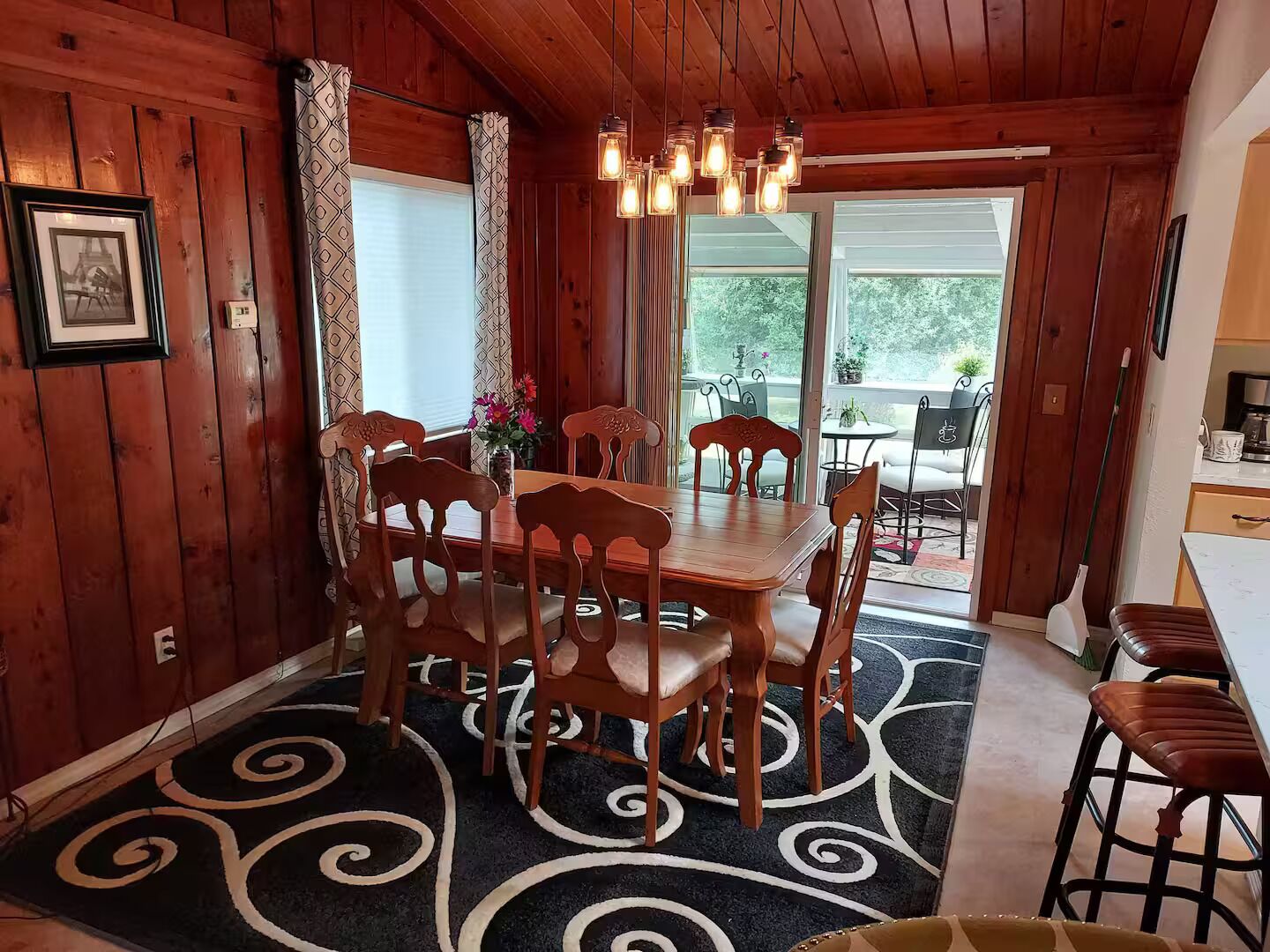A wooden-paneled room with a dining table and chairs, a chandelier, and a patterned rug.