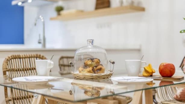 Ein Frühstückstisch mit Korbstühlen, einer Glasebene, einer Auswahl an Backwaren, einer weißen Tasse, einem gelben Servietten und einem roten Apfel.