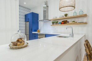 Eine moderne Küche mit weißer Arbeitsplatte, blauen Schränken und einer Edelstahl-Dunstabzugshaube. Darüber befinden sich Holzregale mit verschiedenen Küchenutensilien.