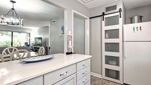 Una cocina con encimera blanca, comedor con mesa y sillas, y puerta corrediza de vidrio que da a otra habitación.