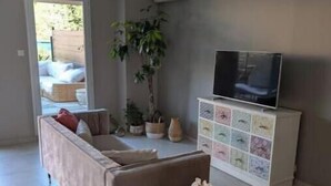 Una sala de estar con un sofá, un televisor en un mueble bajo, una planta y una mesita con un florero.