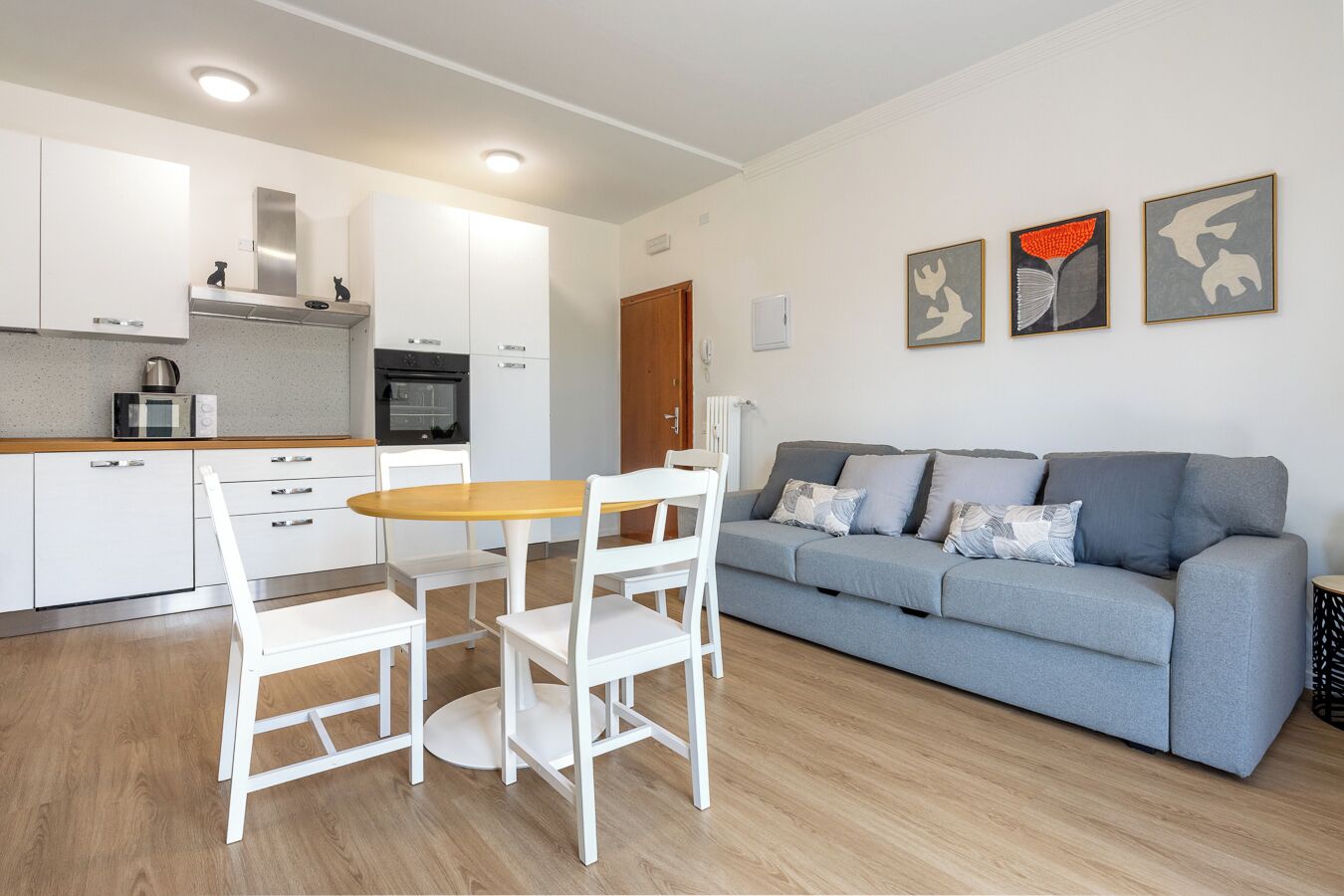 A modern kitchen with white cabinets, a wooden table, and a grey sofa in a living area.
