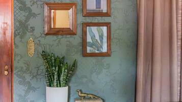 Een kamer met behang met patroon, een houten kast met lades, een potplant en ingelijste foto's aan de muur.