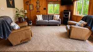 Ein gemütliches Wohnzimmer mit grauem Sofa, zwei braunen Ledersesseln, einer Holzwand mit Hirsch- und Fischdekor, einem Fernseher und einem Fenster mit Blick auf einen Wald.