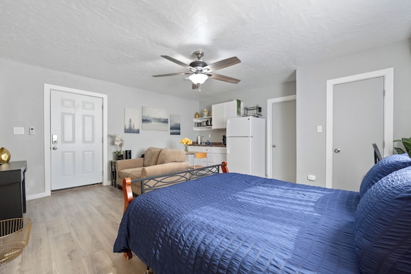 A bedroom with a bed, a ceiling fan, a kitchenette, and a door.