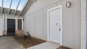 Un bâtiment à la façade blanche, une porte ornée d’un panneau « interdiction de fumer », une petite marche menant à l’entrée et un espace abrité avec une lucarne.