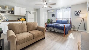 Un salon moderne comprenant un canapé, un lit avec un cadre en métal, un ventilateur de plafond et un coin cuisine avec des placards et un plan de travail.