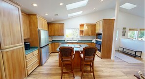 A modern kitchen with wooden cabinets, a central island, two bar stools, and a view of the outdoors. - Amazing Ocean view 1 mile to Brookings! (Brookings)