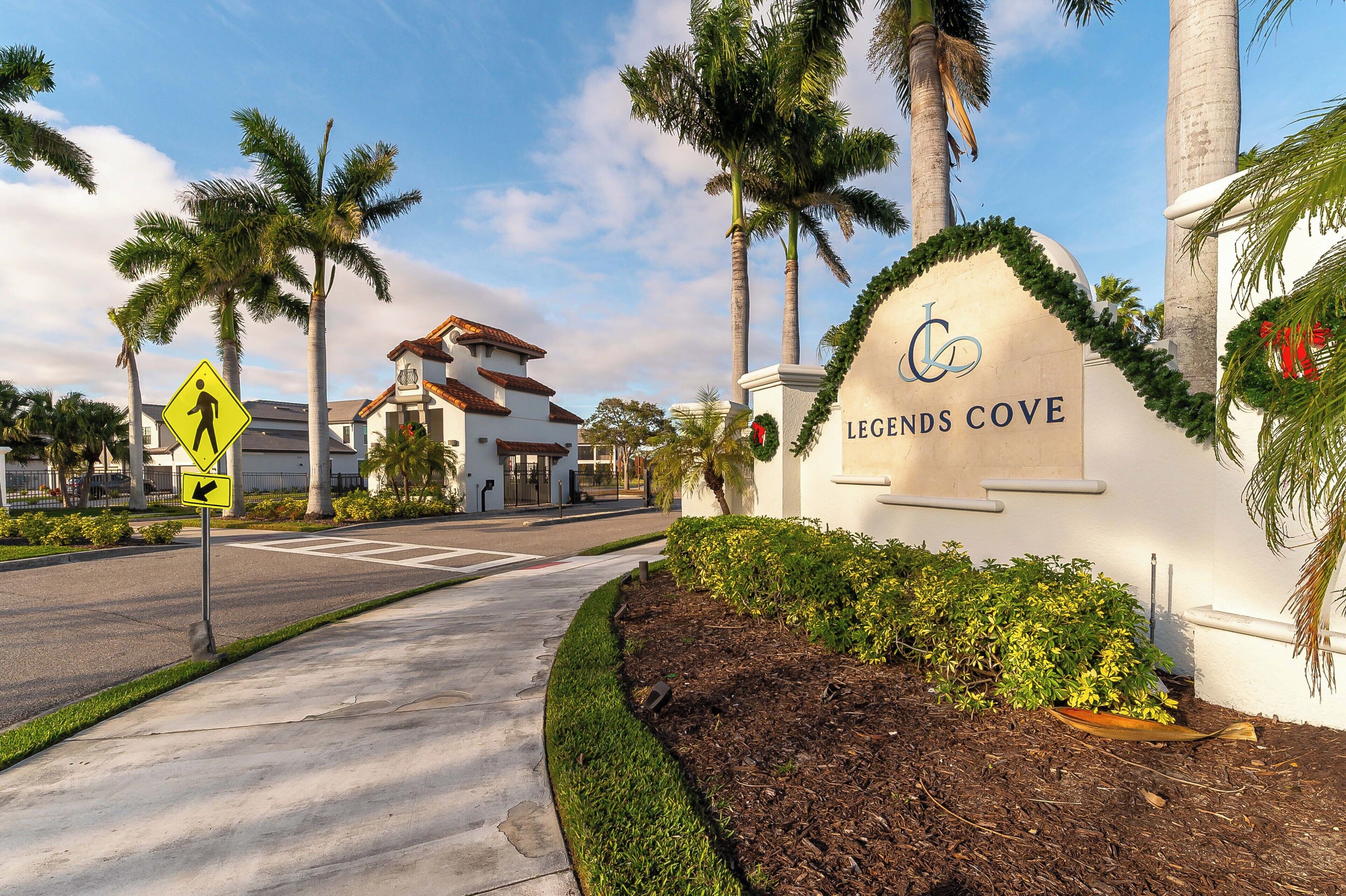 A sign for Legends Cove with a pedestrian crossing sign, palm trees, and a building with a tiled roof.