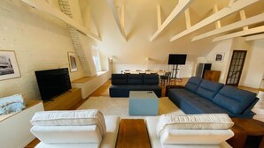 A spacious living room with a high ceiling, a dark blue sofa, white armchairs, a wooden coffee table, and a television. - Les Ancestrales - Maison Turcot Art Déco (Saint-Hyacinthe)