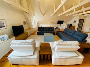 A spacious living room with a high ceiling, a dark blue sofa, white armchairs, a wooden coffee table, and a television.