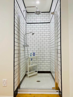 A small, enclosed shower area with white and black tiled walls, a white shelf with toiletries, and a visible drain. - Les Ancestrales - Maison Turcot Art Déco (Saint-Hyacinthe)