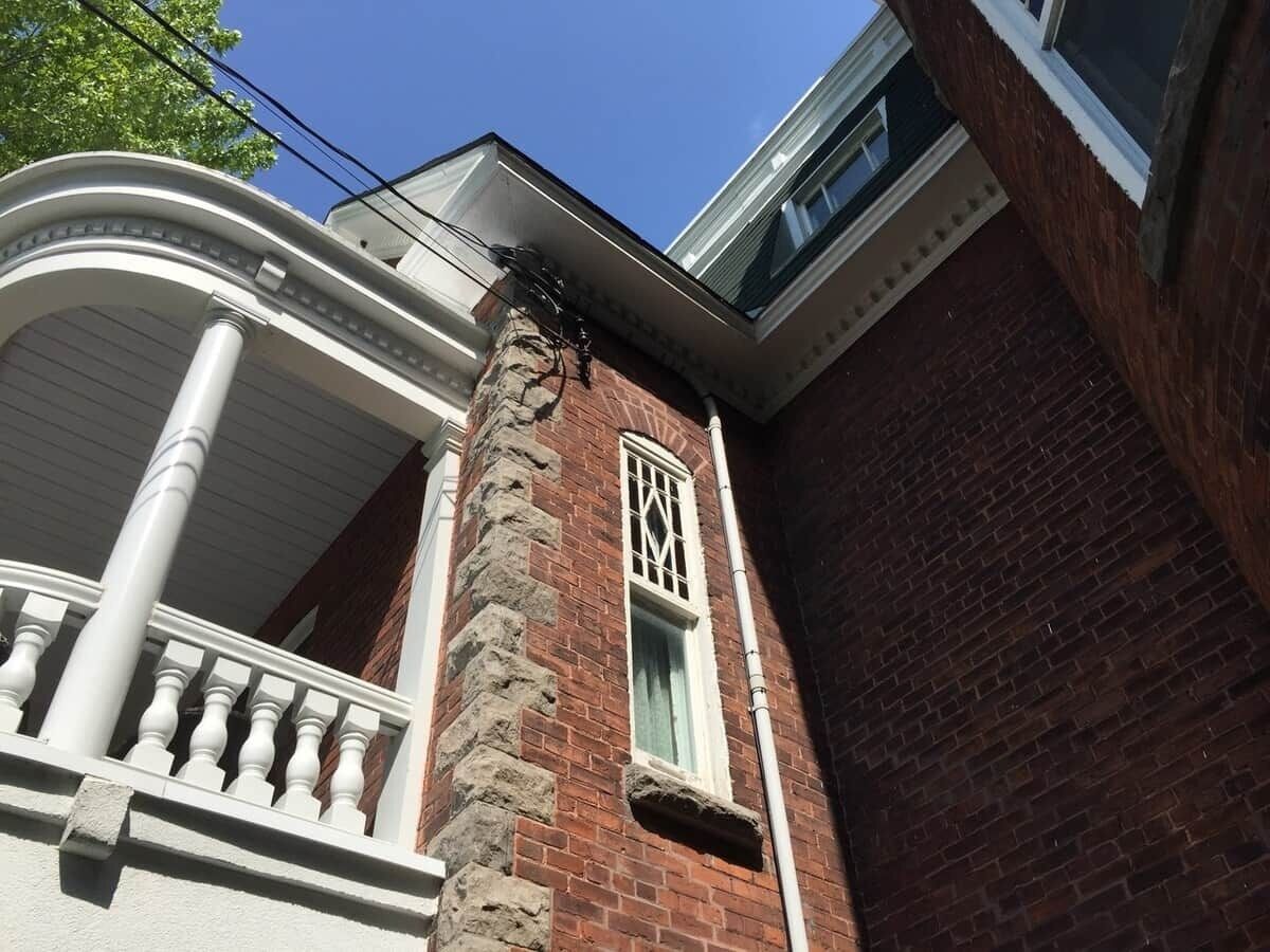 Een hoek van een gebouw met een bakstenen gevel, een wit balkon met balustrades en een raam met decoratief roosterwerk.