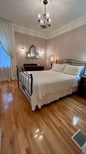 A bedroom with a bed, a nightstand, a dresser, a mirror, a chandelier, and a window with curtains.
