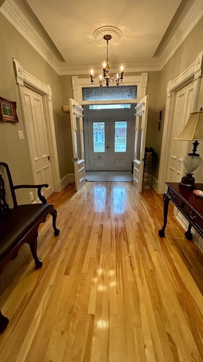 A hallway with wooden flooring, a chandelier, and double doors leading to a room.