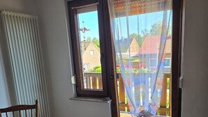 A room with a dining table, chairs, a radiator, a sliding glass door with a view of houses, and a trash bin. - Vacation apartment in Emsland (Papenburg)