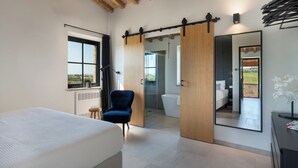 Un dormitori modern amb un llit gran, una cadira, una porta corredissa que condueix a un bany i un mirall que reflecteix l'habitació.