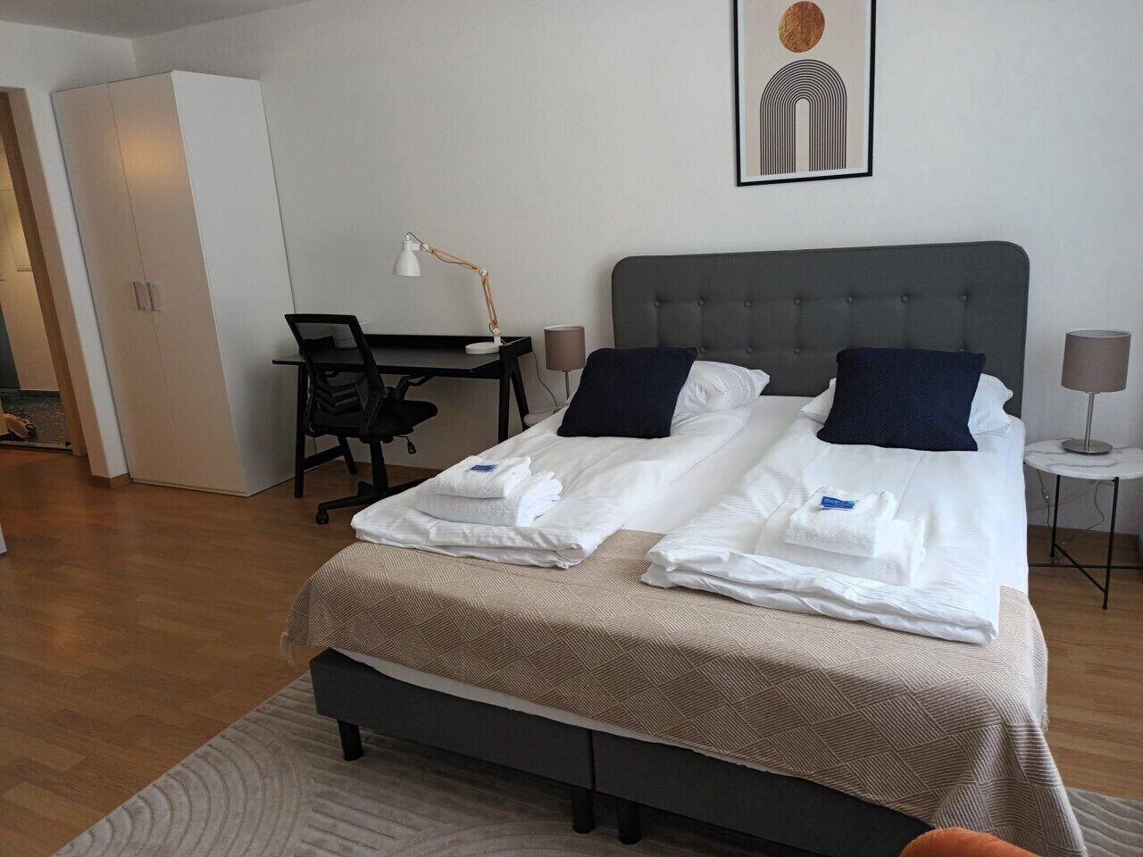 A neatly made bed with pillows, a desk with a chair, a lamp, and a framed picture on the wall.