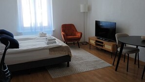 Hotellihuone, jossa on sänky, tuoli, työpöytä, televisio ja ikkuna, jossa on verhot.