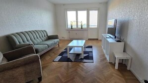 A living room with a sofa, a coffee table, a TV stand, and a window with a view. - Cozy apartment near Volvo Cars (Gothenburg)