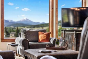 Une pièce comprenant un canapé, un téléviseur, une table avec une bouteille et un verre, et une fenêtre donnant sur la montagne.
