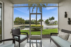A room with a glass door leading to a view of a water body, palm trees, and a grassy area. There are two wicker chairs with cushions. - 814 - Lake and Fountain View - Chic Furnishings (Naples)