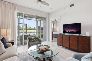 Ein modernes Wohnzimmer mit einem Glastisch, einer Couch, einem Fernseher auf einem Holzkasten und einer Glastür, die zu einem Außenbereich führt.