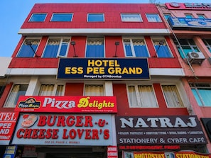 Un immeuble à plusieurs étages, sur lequel on peut voir des enseignes indiquant : Hôtel Ess Pee Grand, Pizza Delights, Burger & Cheese Lover's et Natraj Stationery & Cyber Café.