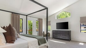 A bedroom with a four-poster bed, a flat-screen TV, a dresser, and a view of greenery through the windows. - 6 BR Mountain Golf Retreat, Hot Tub, Fall Foliage Views, Trailhead Steps Away (Avon)
