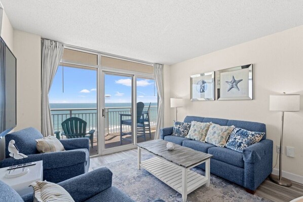 A living room with a blue sofa, a coffee table, and a view of the ocean through a sliding glass door. - Oceanfront Condo with Lazy River (North Myrtle Beach)