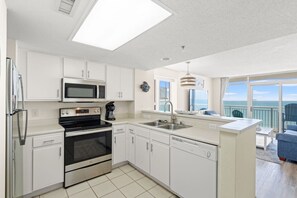 A modern kitchen with white cabinetry, a microwave, stove, and dishwasher, and a balcony with a view of the ocean. - Oceanfront Condo with Lazy River (North Myrtle Beach)