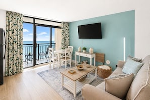 Ein Wohnzimmer mit Sofa, Couchtisch, Fernseher, Esstisch mit Stühlen und einem Balkon mit Blick.