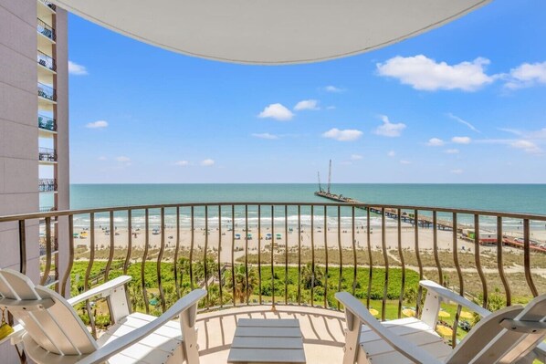 Ein Balkon mit weißen Stühlen und Blick auf einen Strand mit einem Steg.