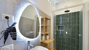 Une salle de bain moderne avec un grand miroir ovale, un lavabo blanc, une étagère en bois, une douche carrelée de carreaux verts et un porte-serviettes.