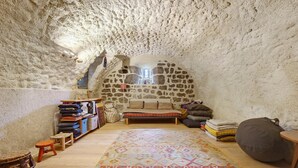 Un interno con una parete in pietra, un letto, una panca, un tappeto e uno sgabello.