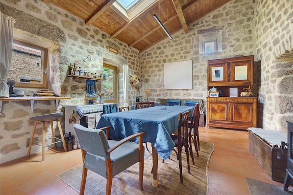 Une piĂšce aux murs de pierre et au plafond en bois, avec une table et des chaises, une fenĂȘtre et un meuble sur lequel sont posĂ©s divers objets.