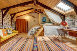 A rustic room with a stone wall, wooden beams, a bed with a canopy, a desk, and a colorful rug.