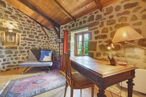 Une piĂšce rustique avec des murs en pierre, un plafond en bois, un bureau avec une lampe, une chaise, un canapĂ© et une fenĂȘtre avec des rideaux.
