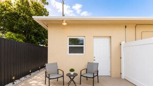 Een gelijkvloers gebouw met een beige gevel, een witte deur en een klein raam met zonwering. Er staan twee stoelen met kussens en een kleine tafel met een bloempot.